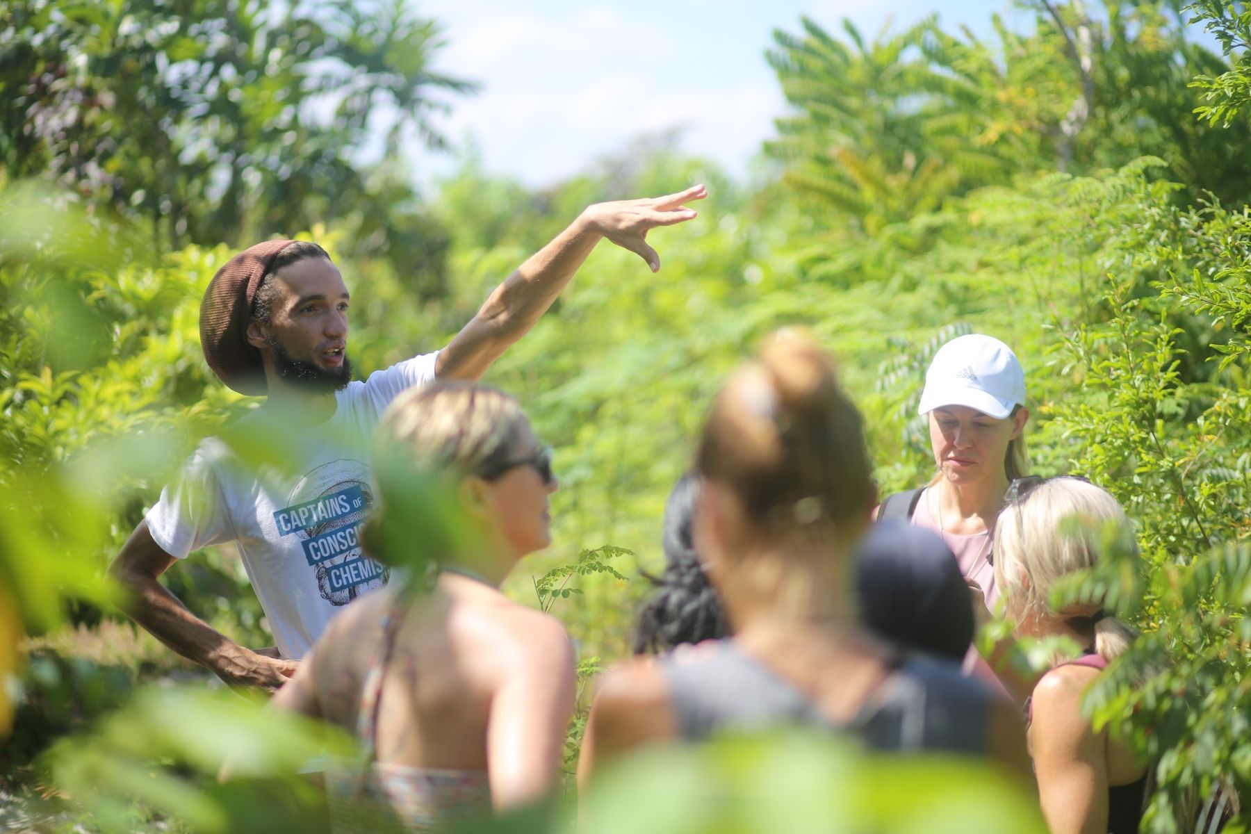 A local guide gesturing toward lush green growth as a group listens during a farm tour.