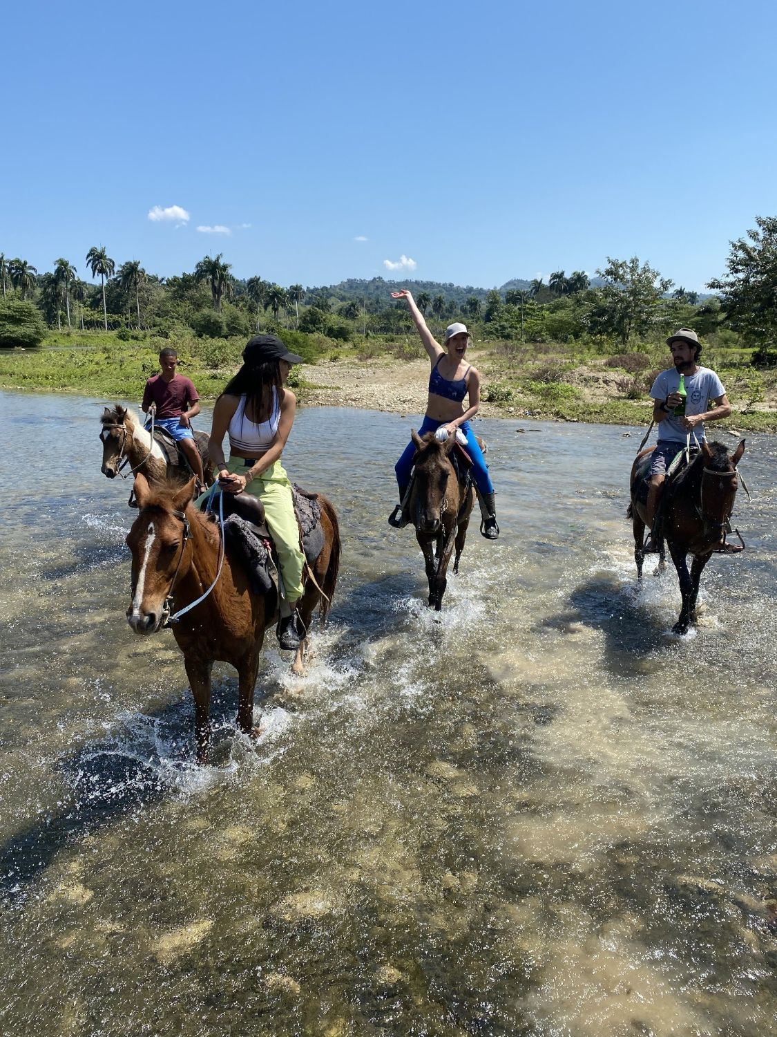 Riders on horseback crossing a shallow river, one raising an arm, with palms and green hills behind.