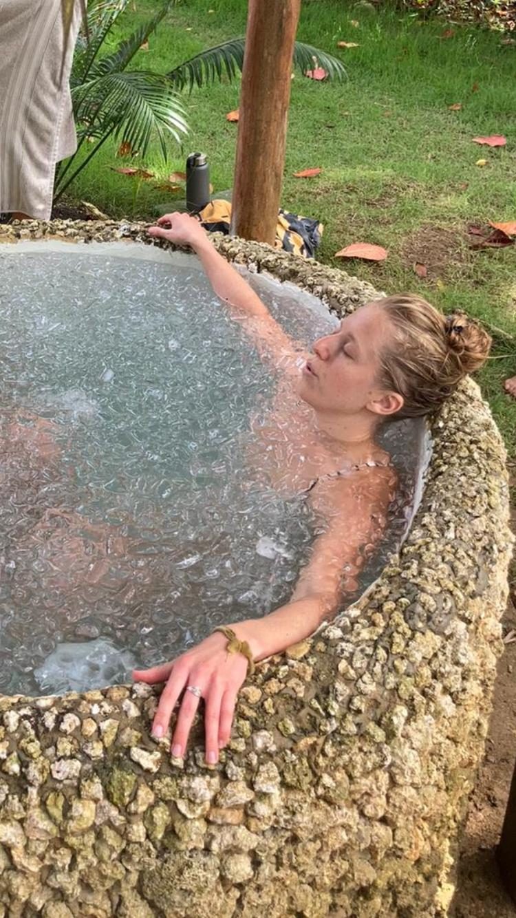 Eine Person ruht in einem Eisbad im Freien, in einem runden Steinbecken, umgeben von Gras.