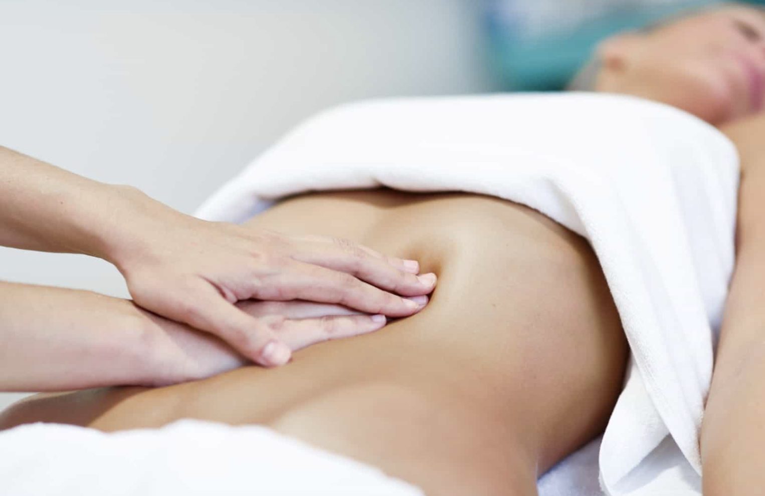 Hände massieren den Bauch einer Person, über die ein weißes Handtuch gelegt ist.