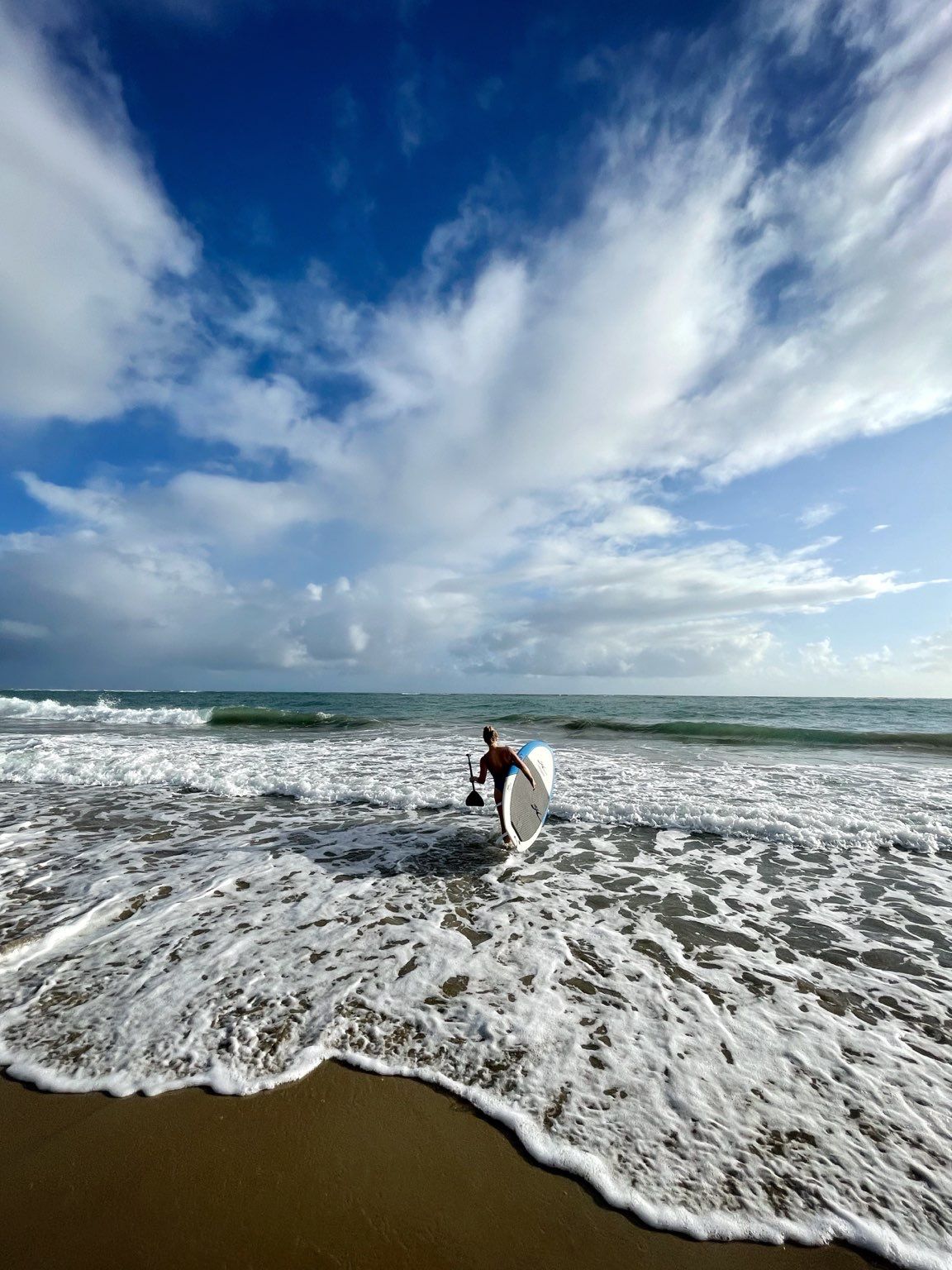 A paddleboarder carrying a board and paddle into the surf beneath a huge cloudy sky.
