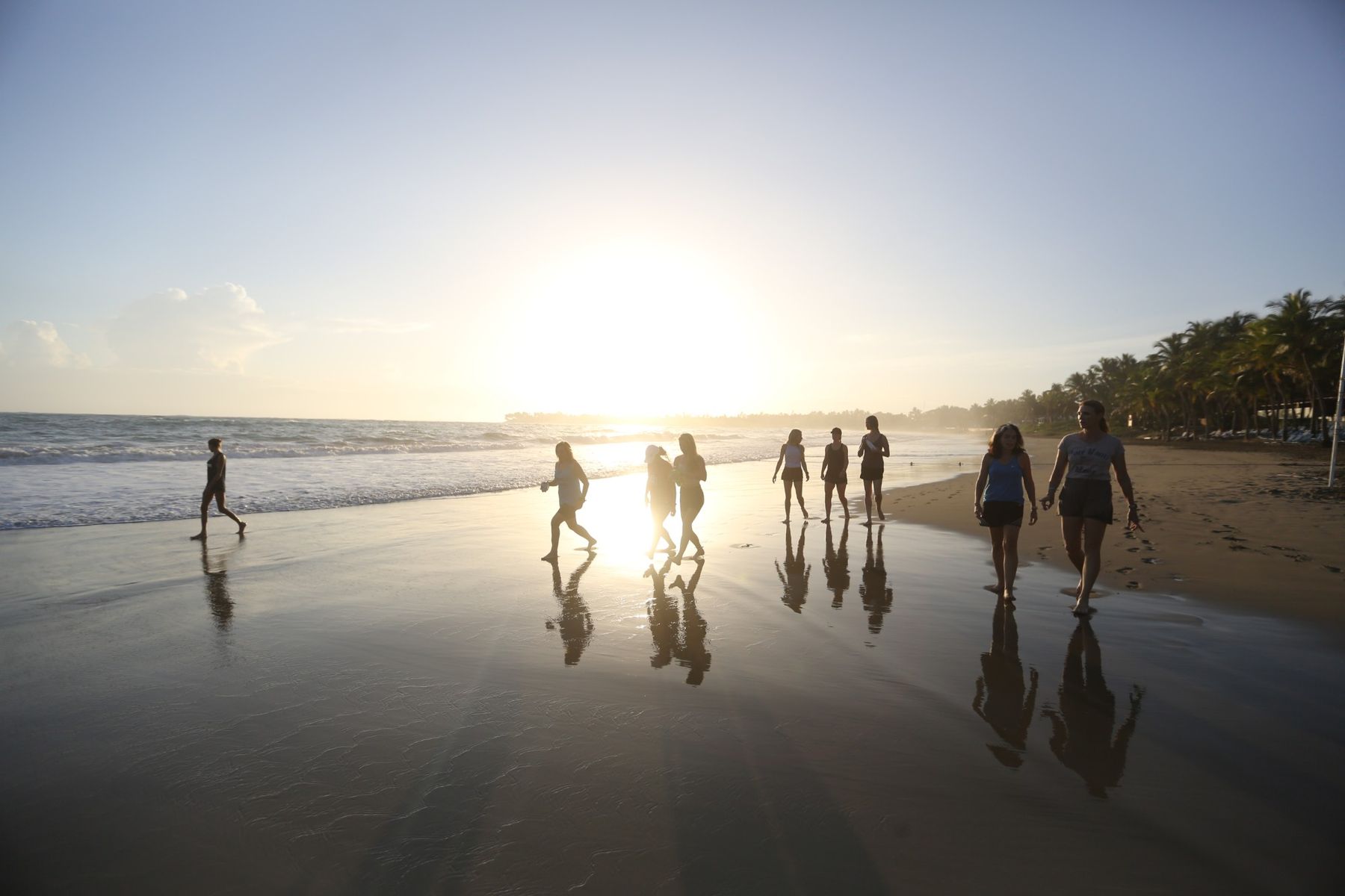 A group of people in silhouette walking along a wet beach at sunrise, their reflections on the sand.