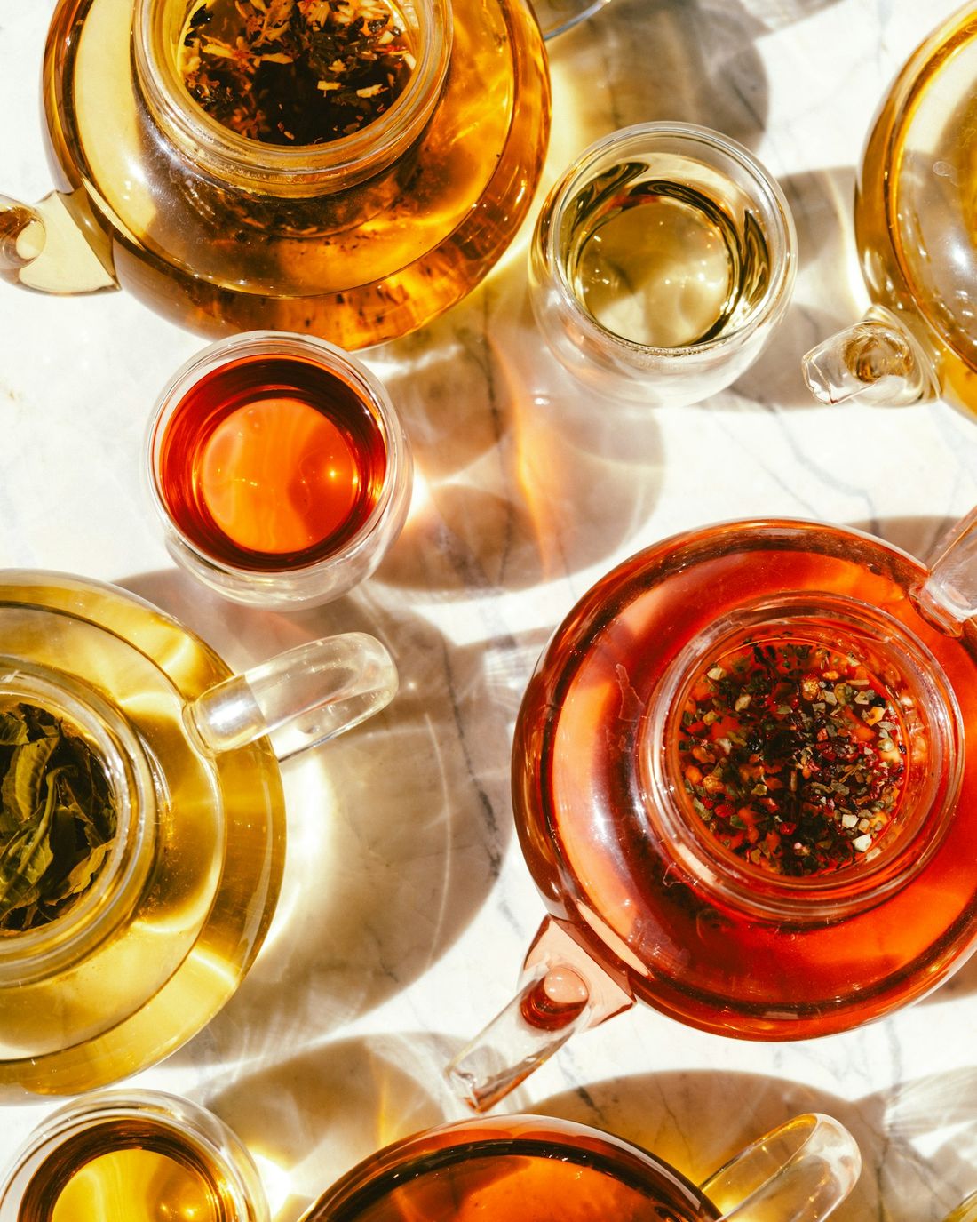 Teteras y tazas de vidrio con té de hierbas y té negro bajo una luz cálida.